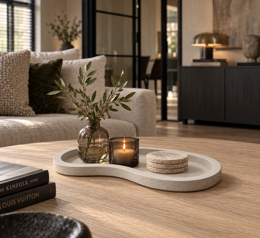 Organisch gevormd decoratief dienblad met vaas, kaars en onderzetters op een houten salontafel in een warm modern interieur.